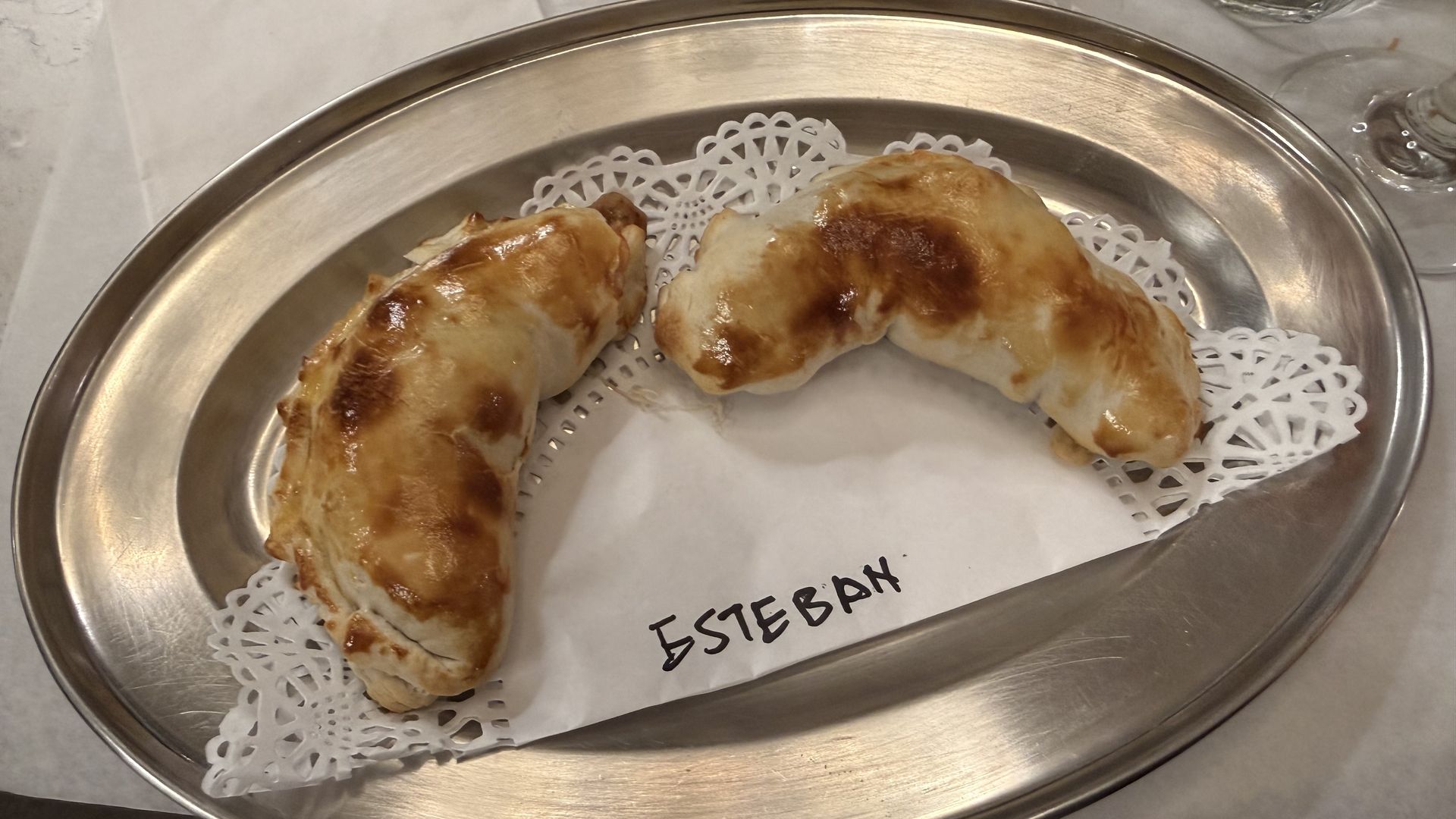 Two golden-brown baked empanadas on a silver tray with a white sheet and a paper labeled "ESTEBAN." 