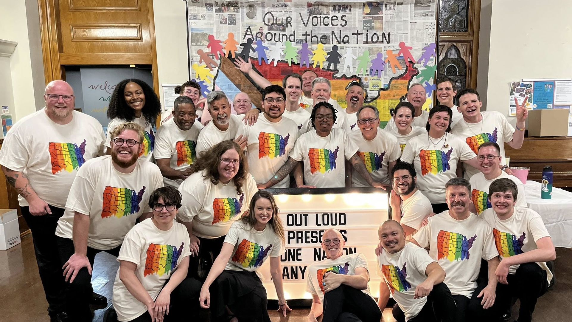 Two dozen men wearing rainbow shirts pose for a photo, smiling. 