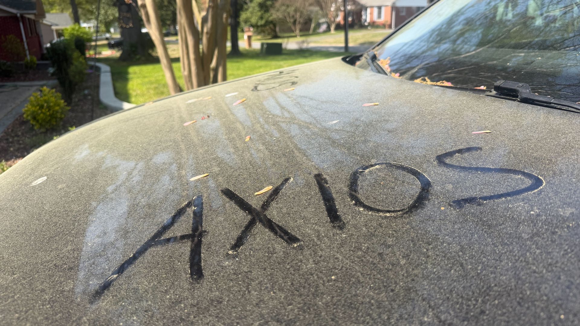 Dusty car hood with the word "AXIOS" drawn in dark marker; scattered leaves, windshield wipers, and a suburban street with houses and trees in the background.