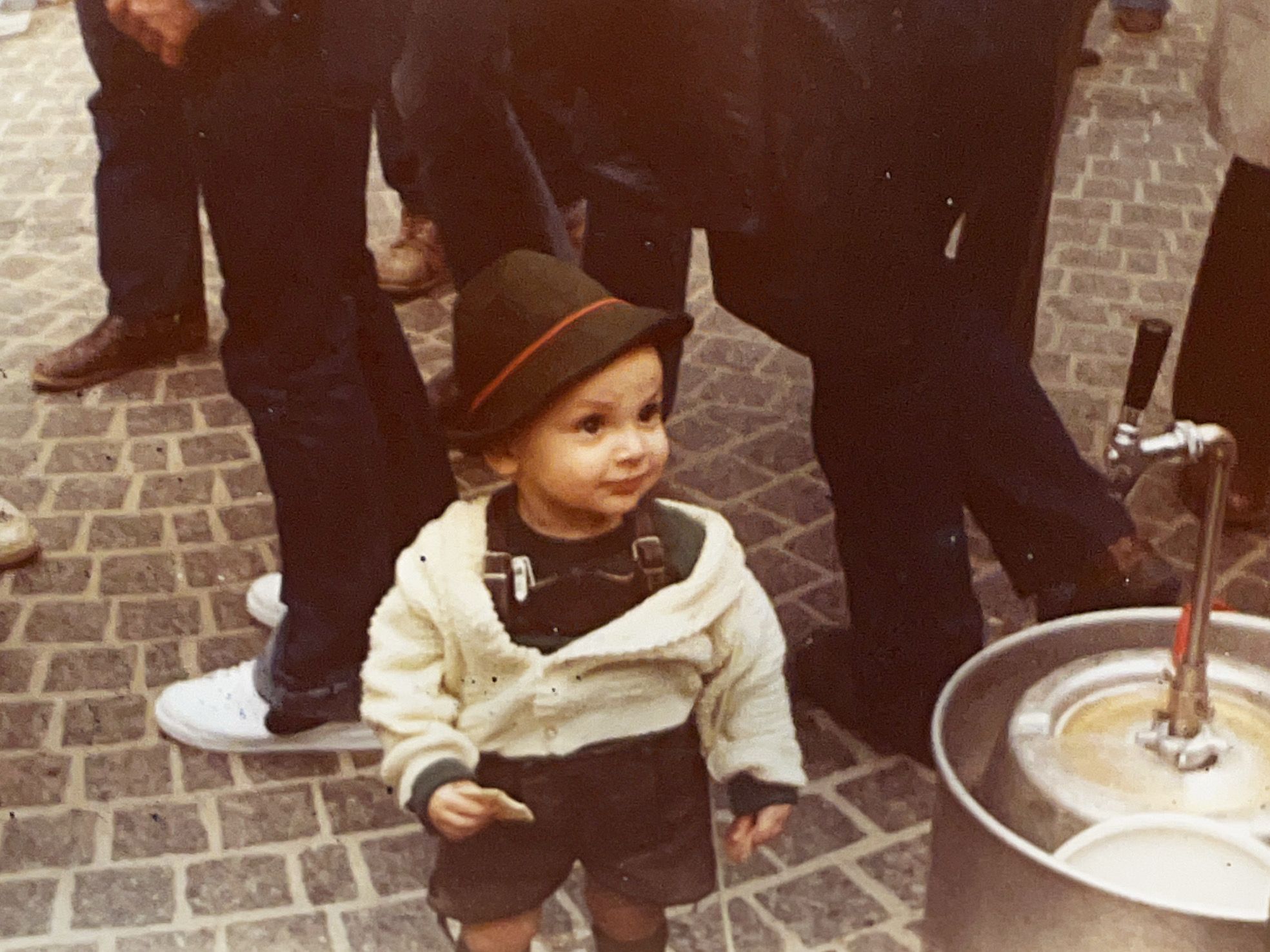 A young child in a cream fleece jacket and brown hat with orange trim stands on a gray brick-paved street, surrounded by adults' legs; a stroller is nearby.