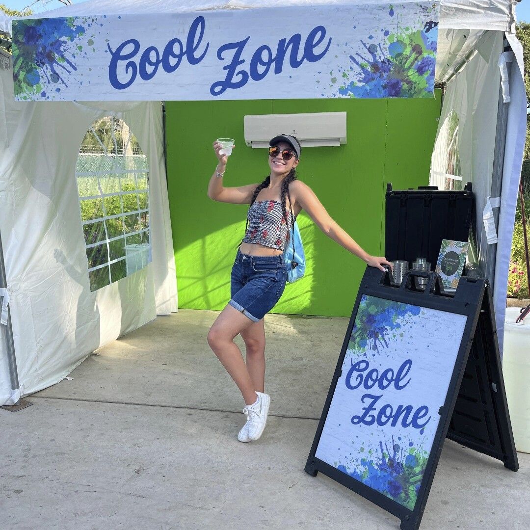 A woman poses with a cup of water at a small tent labeled "cool zone."