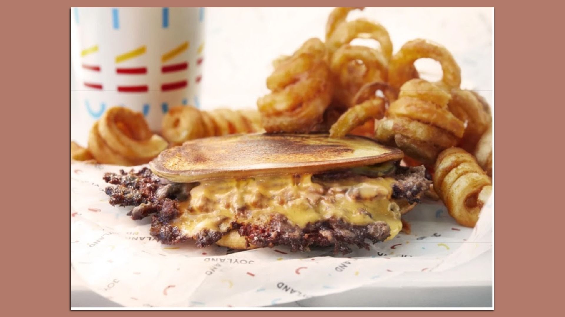 Close-up of a cheeseburger with melted cheese and a toasted bun, surrounded by golden onion rings and curly fries; a colorful striped cup is in the background.