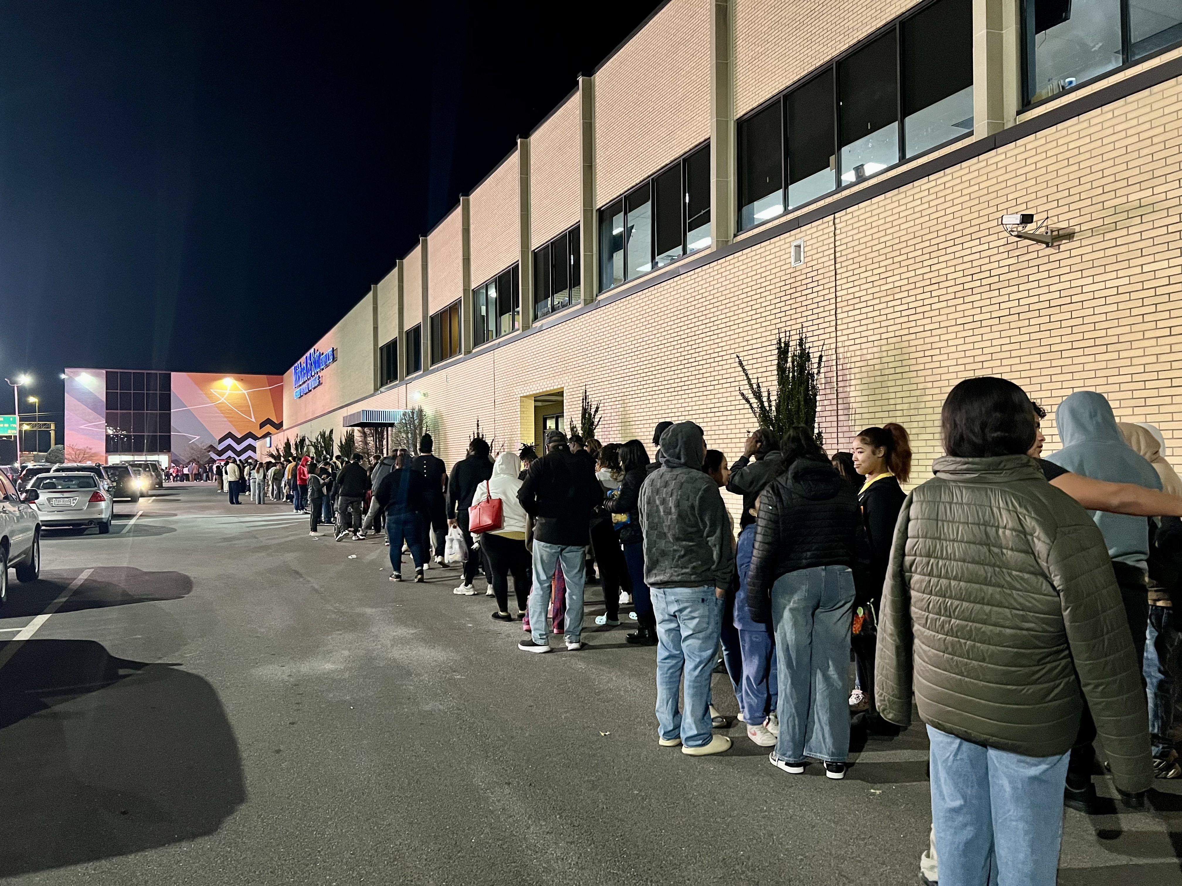 Long line of people in jackets and hoodies waiting outside a large beige brick building at night, with cars parked nearby and a mural visible in the distance.