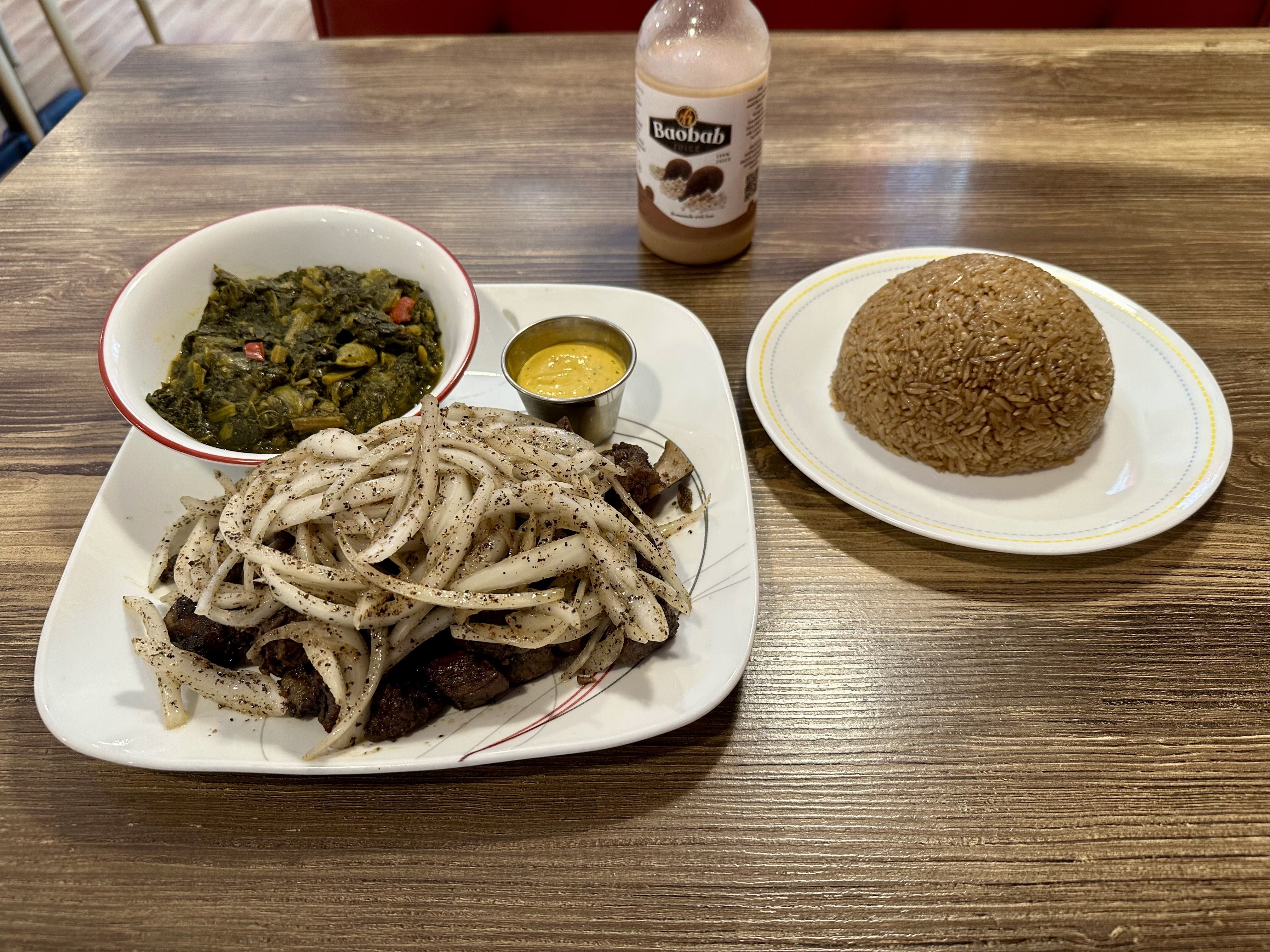 Meal on a wooden table: pepper-speckled flat noodles, a greens dish with peppers and a small yellow dipping sauce, a mound of brown rice on a separate plate, and a Baobab juice bottle in the background.