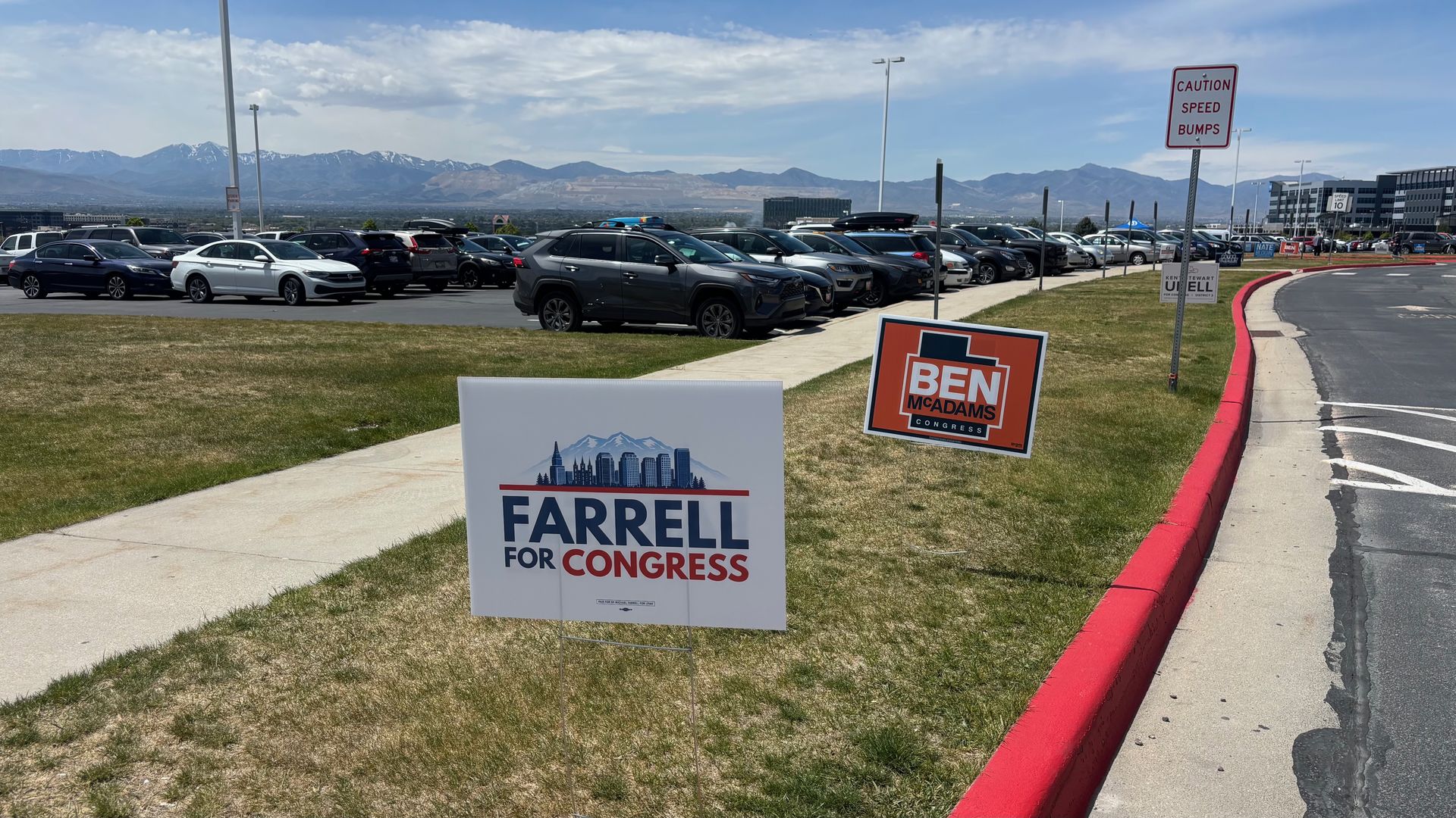 Wide parking lot full of cars with campaign signs for Farrell for Congress and Ben McAdams; grassy foreground, red curb, distant office buildings and snow-capped mountains under a blue sky.