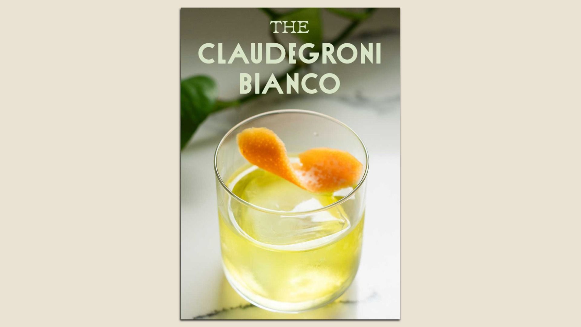 A glass of pale yellow cocktail with a large ice cube and orange peel garnish, labeled as "The Claudegrobi Bianco" on a light marble surface with green leaves blurred in the background.