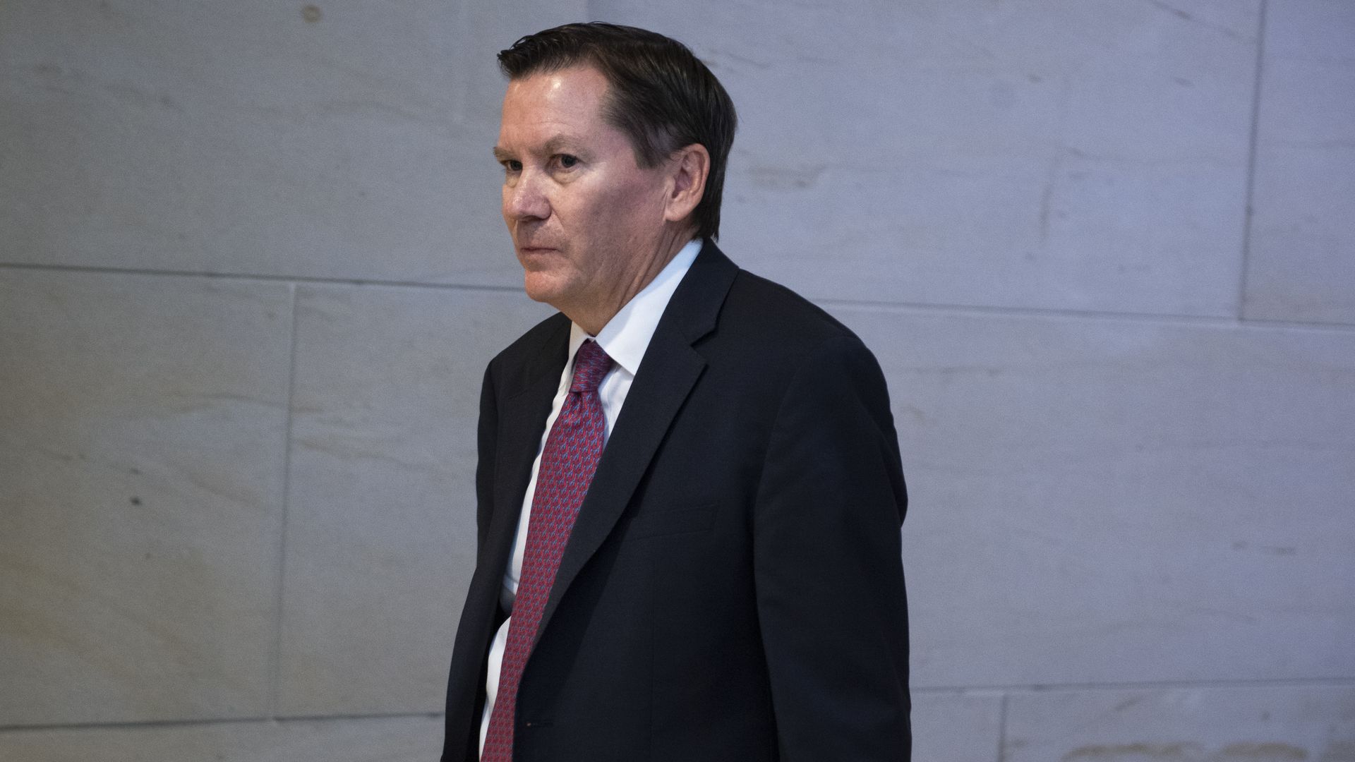 In this image, Atkinson walks down a hallway wearing a suit.