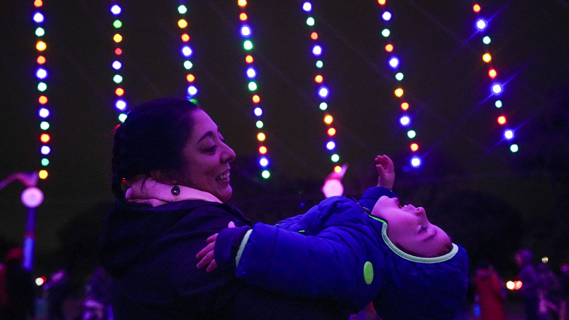 A woman smiling as she holds a child in a blue coat at a nighttime event with colorful vertical string lights in the background and people visible in the distance.