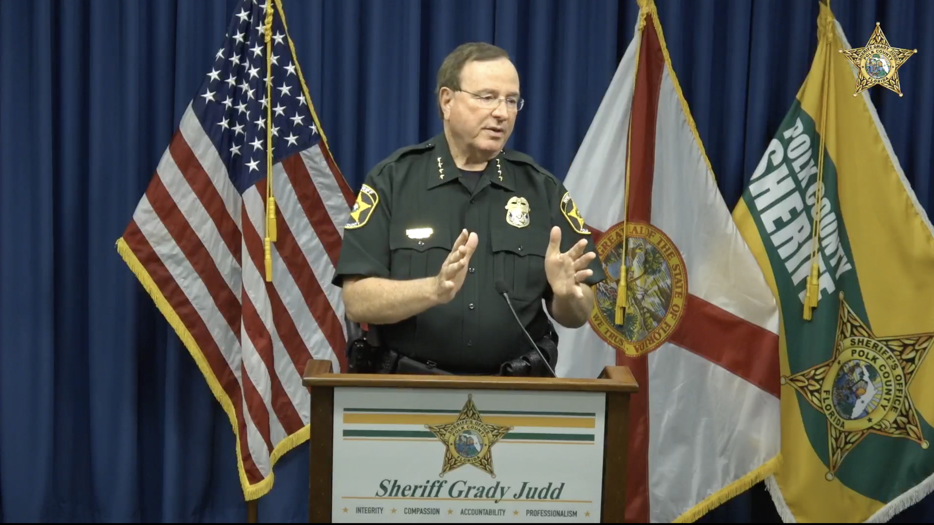Sheriff Grady Judd, in a dark green uniform, speaks at a podium with hands raised. Behind him are the U.S. flag, the Florida flag, and Polk County sheriff banners against a blue curtain.