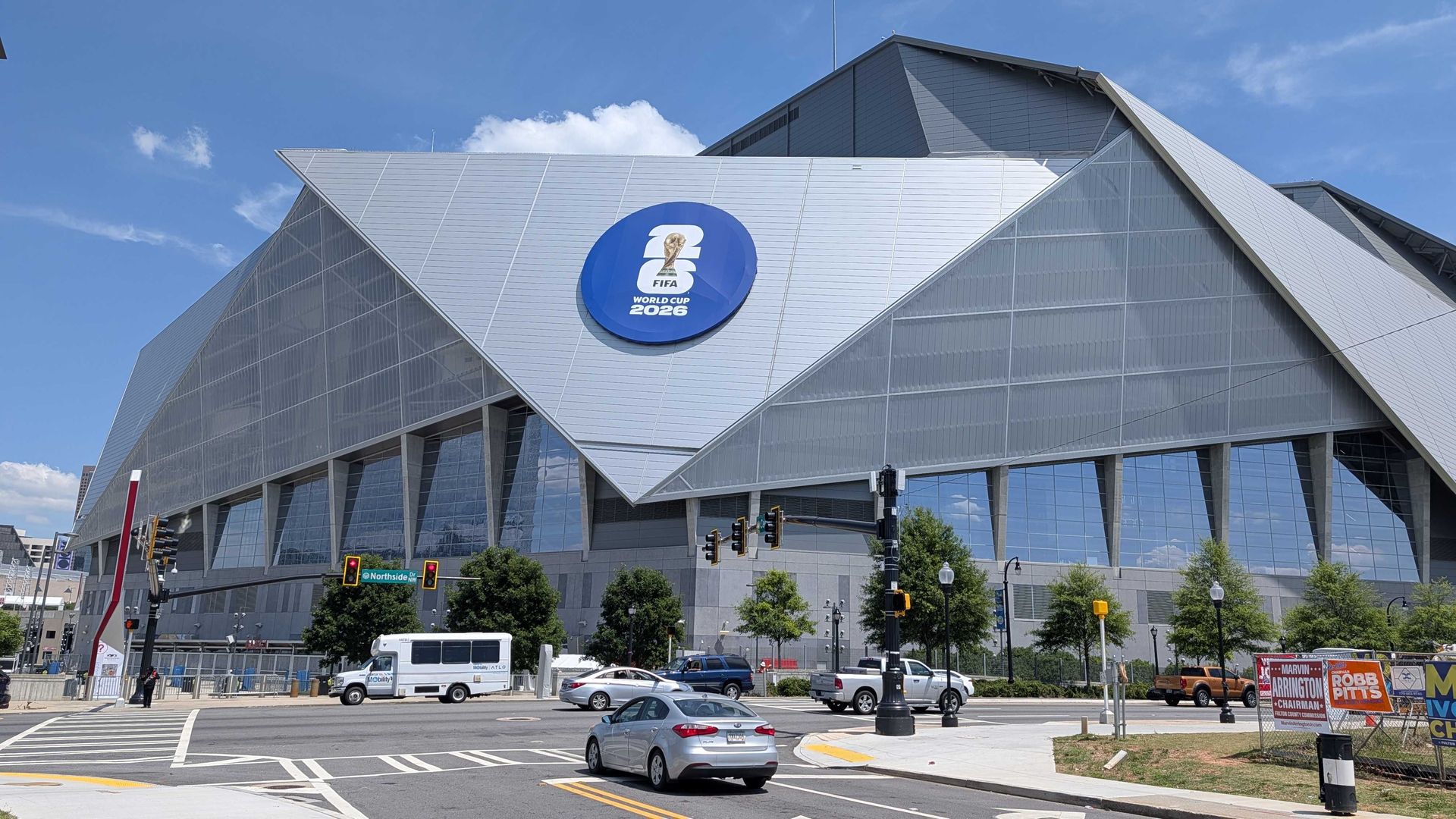 Modern glass-and-metal stadium with a dramatic triangular roof. A large blue circle sign reads "FIFA World Cup 2026" on the facade; cars and traffic in front under a blue sky.