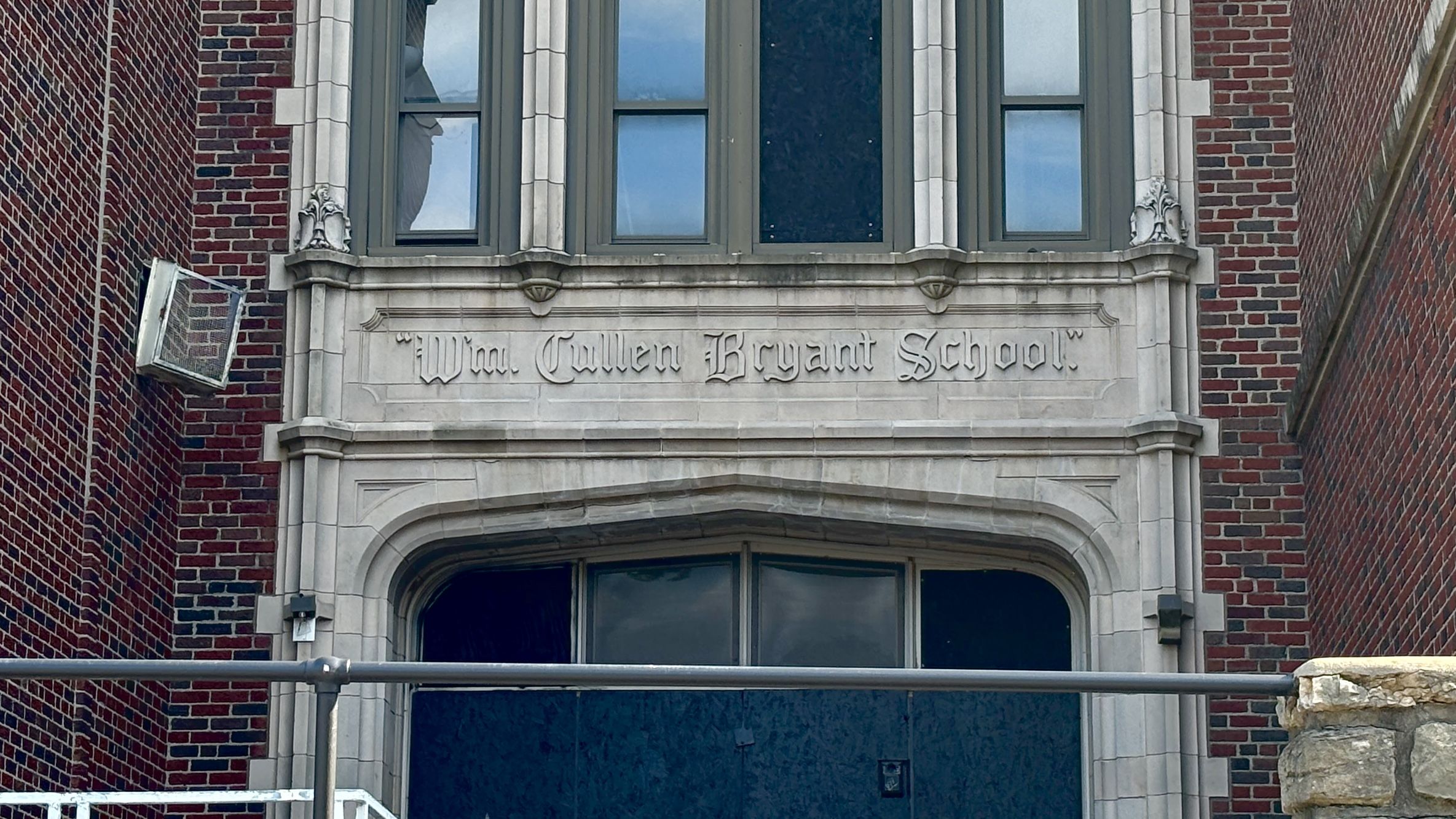 Brick school building facade in red brick with a carved stone plaque reading "Wm. Cullen Bryant School" above an arched entry; flanking stone-trimmed windows and decorative carvings.