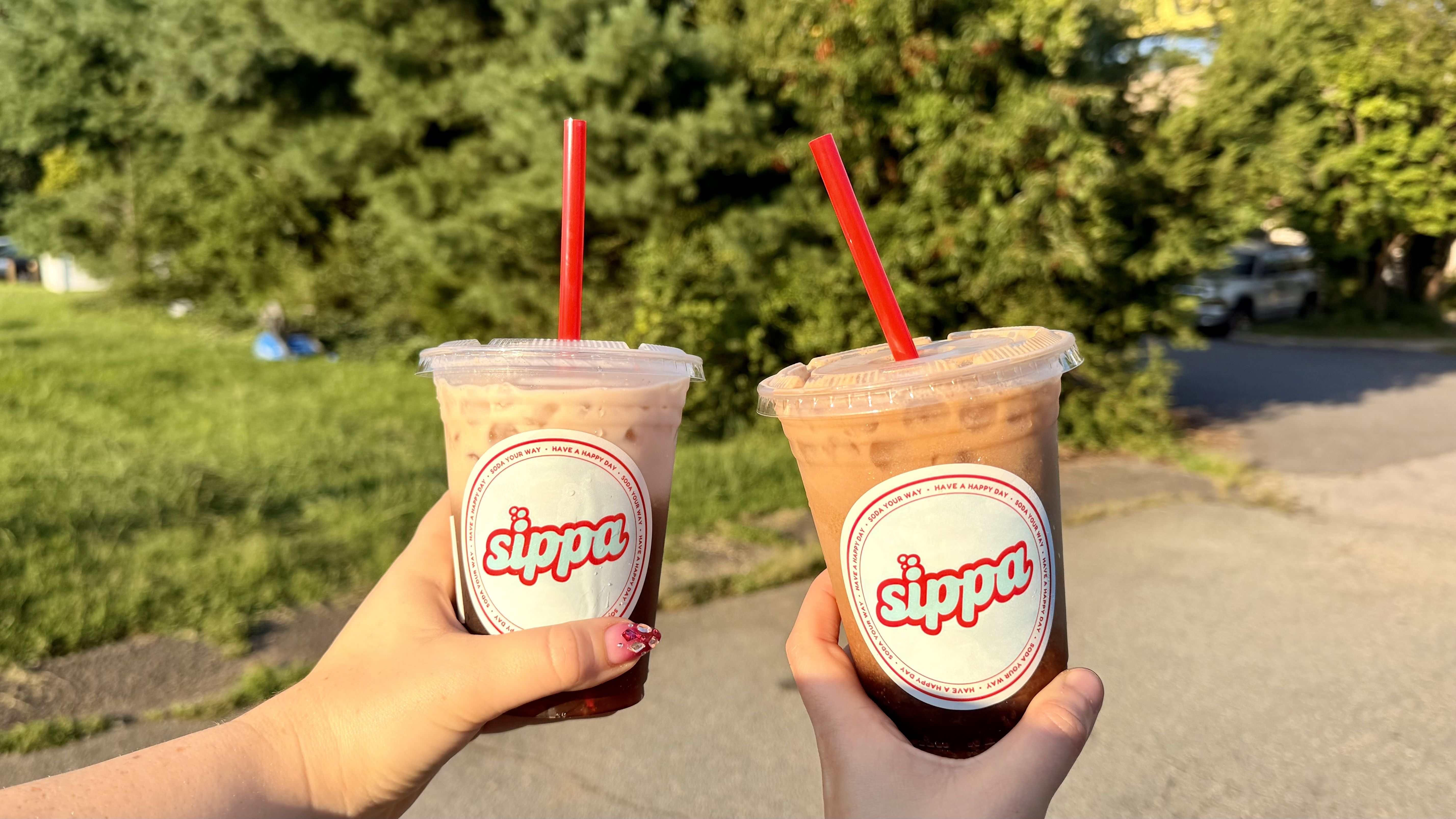 Two iced drinks in clear cups with red straws are raised in front of a sunny park scene; one pink beverage and one brown coffee, hands with nails visible, greenery and a street in the background.