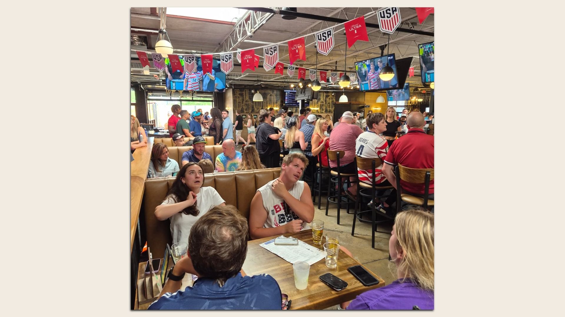 Busy sports bar filled with people seated at tables and standing around the bar; red, white, and blue USA banners hang overhead as multiple TVs show sports highlights.