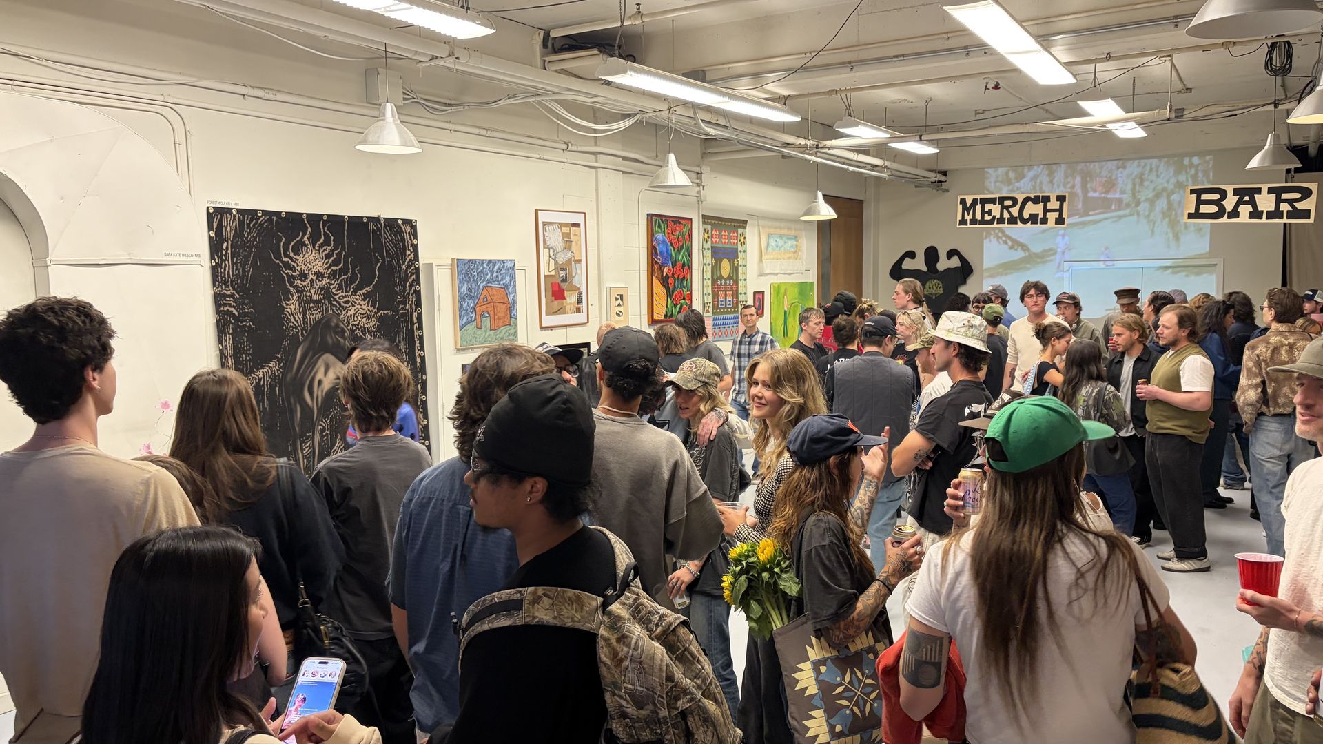 Crowded art gallery with white walls and hanging lights. Attendees mingle, many holding drinks as colorful paintings line the walls. Large MERCH and BAR signs hang above, with a projection in the back.