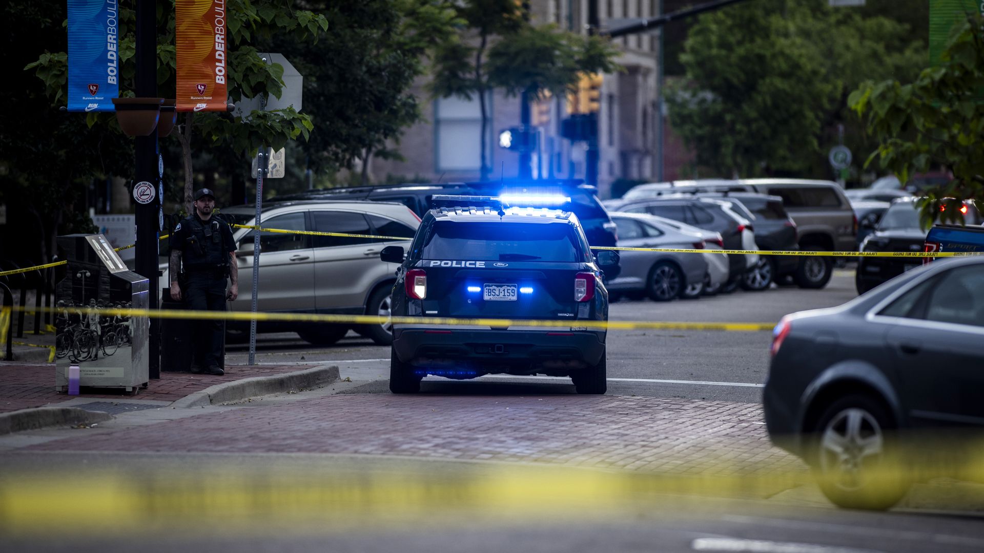 A Boulder police car with flashing blue lights blocks a city street with yellow caution tape, a police officer stands nearby, and parked vehicles line the street.