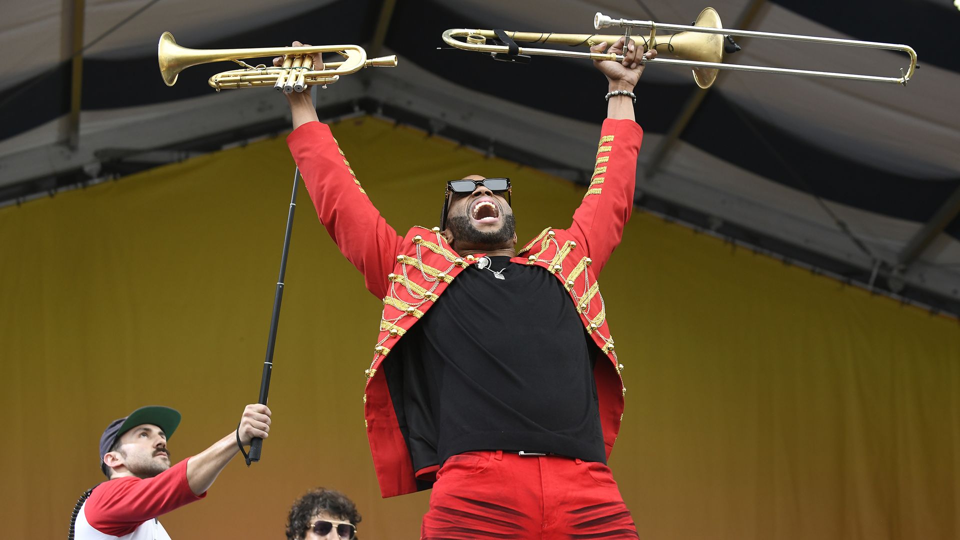 Trombone Shorty lifts his trombone and a trumpet above his head during a performance at Jazz Fest. He yells toward the crowd, unseen.