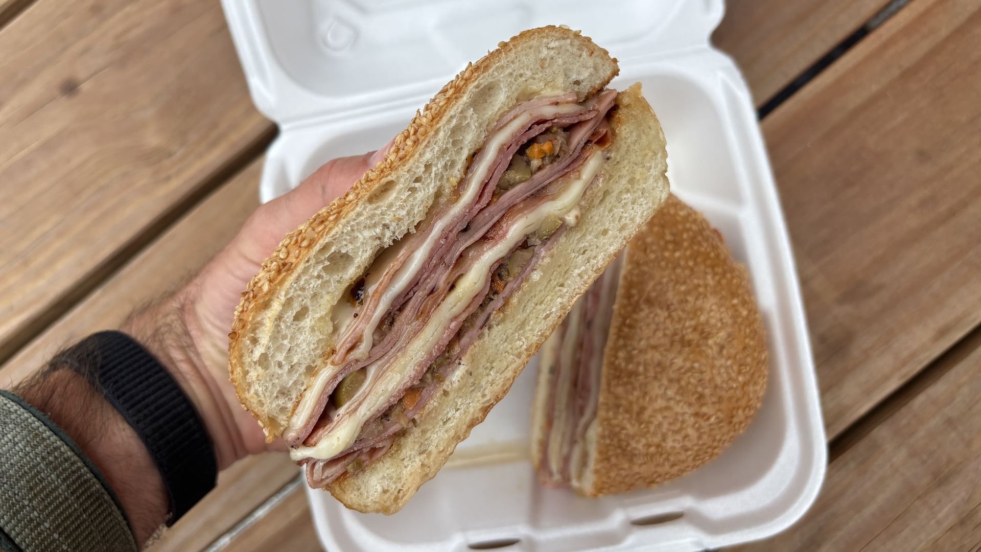 Cross-section of a sesame bagel sandwich with layers of ham and cheese, plus pickles and diced vegetables; held over a white takeout container on a wooden table.