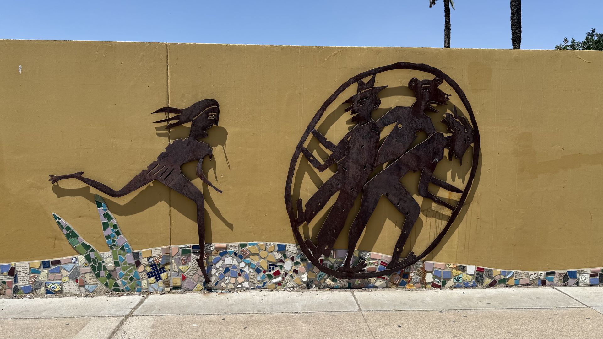 Two-dimensional metal sculptures of a woman and a circle with three human-like figures inside, on a goldenrod colored wall.