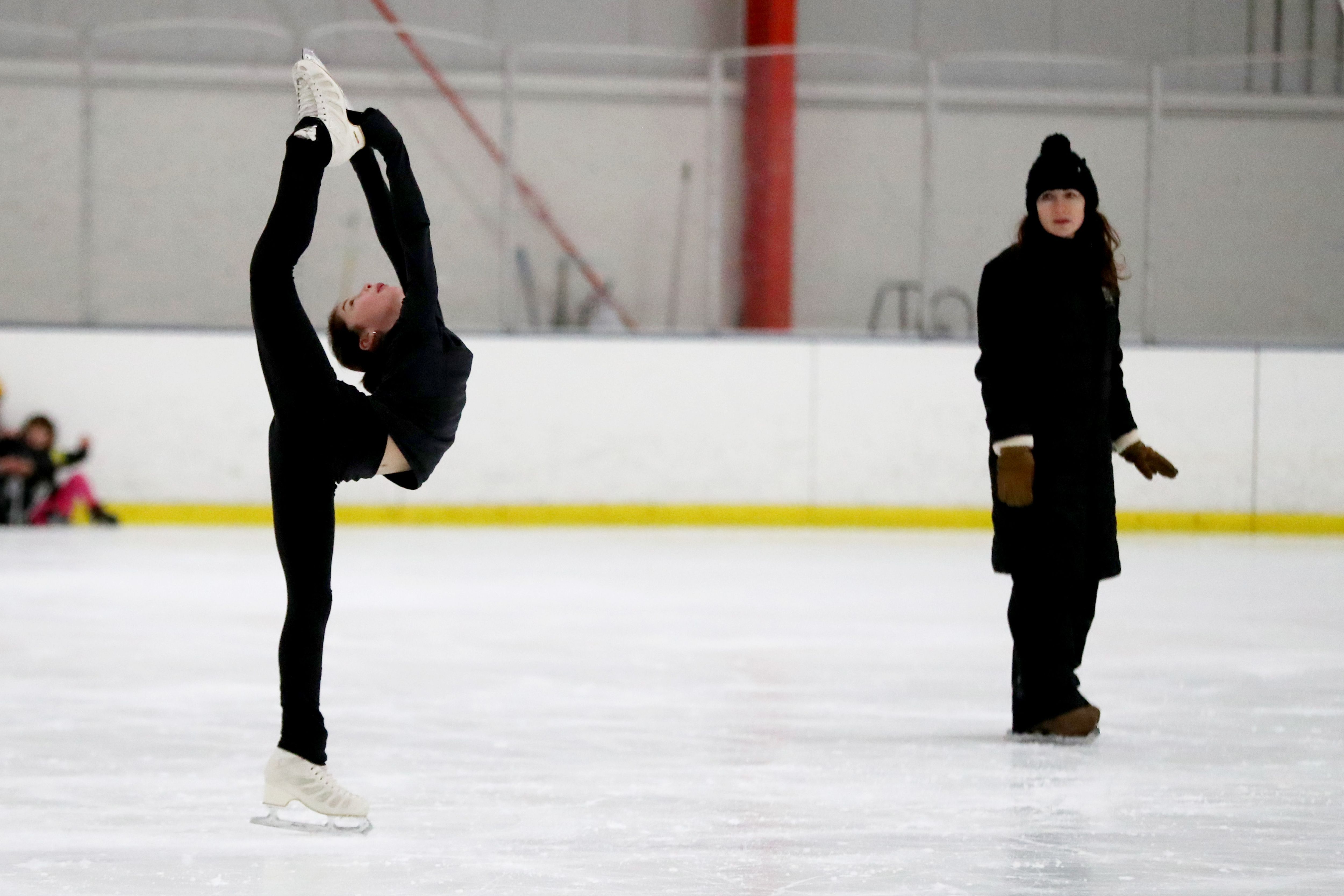 Figure skater in black performs an impressive Biellmann spin on ice, lifting one leg high while balancing on the other. A coach in black winter clothes watches nearby on the rink.
