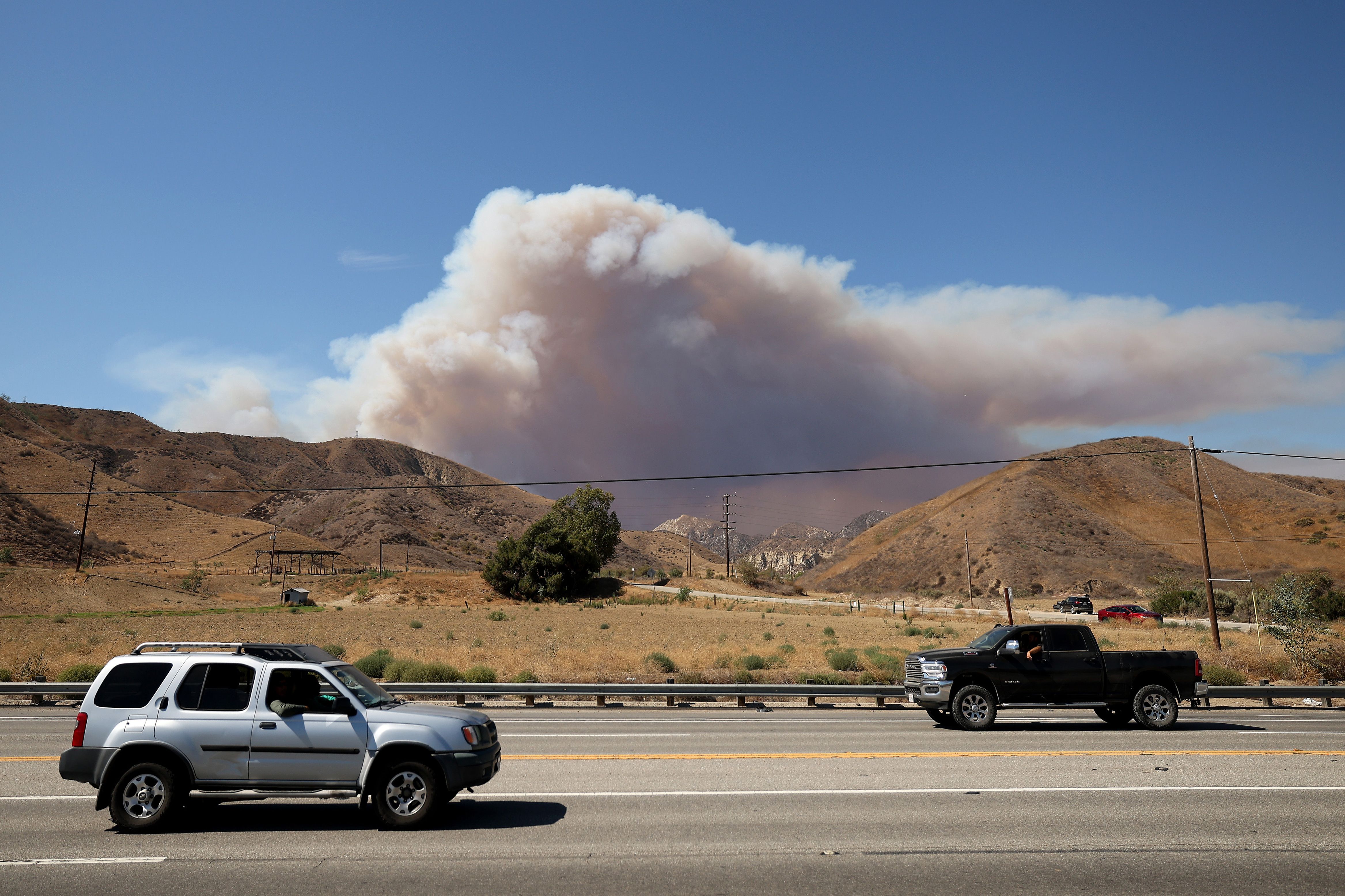 Two vehicles on a road near dry brown hills with a large plume of smoke rising against a clear blue sky, indicating a fire in the distance.