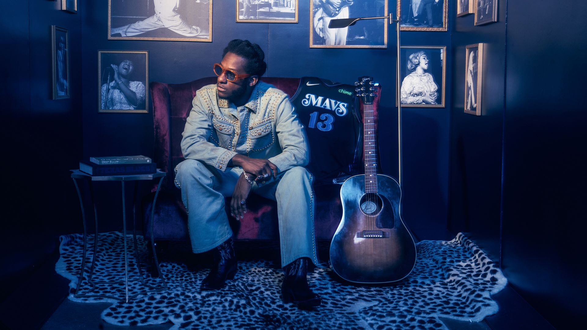 A man sits next to a guitar, surrounded by photos of popular Blues artists.