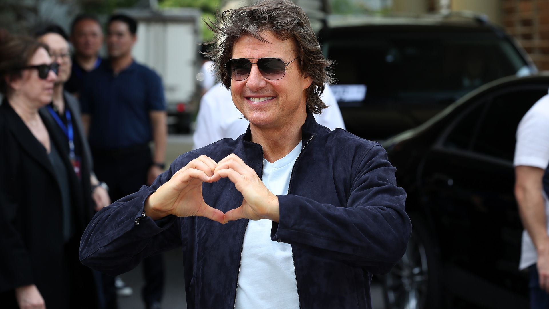 Tom Cruise wearing sunglasses and putting his hands together to make a heart as he greets a crowd