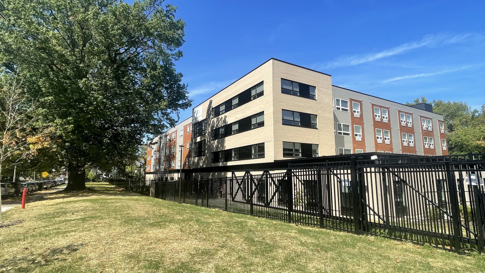 A modern four-story residential building with beige, gray, and red brick walls under a clear blue sky, surrounded by a black metal fence and large green trees on a grassy lawn.