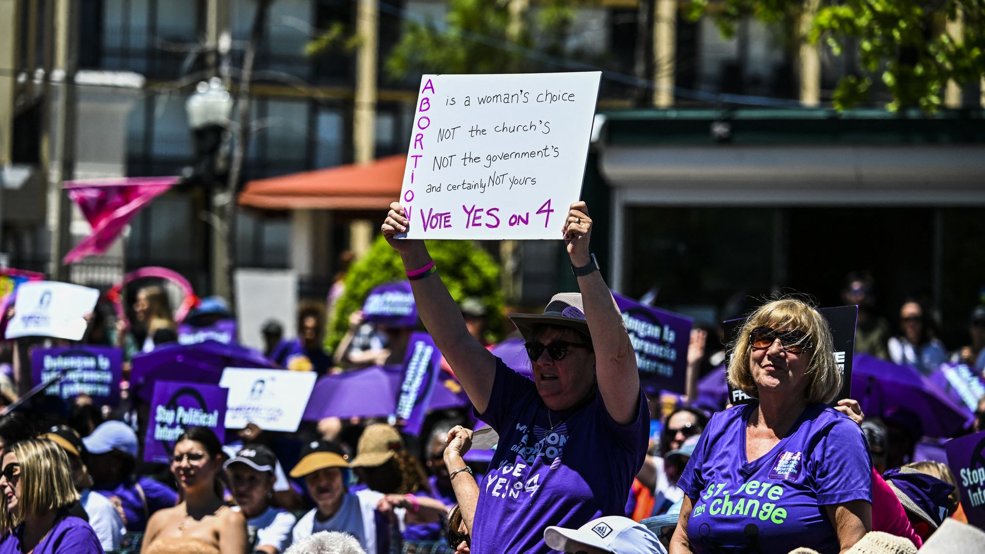 A protester in a purple T-shirt surrounded by other protesters in purple T-shirts holds up a sign that says "Abortion is a woman's choice, NOT the church's, NOT the government's, and certainly NOT yours, VOTE YES ON 4."