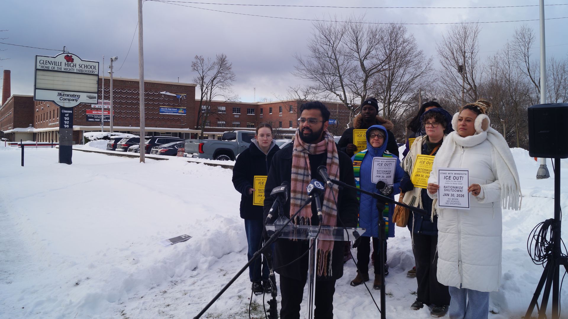 Group of people in winter clothing standing outside snowy Glenville High School, some holding yellow and white signs about a Jan 30, 2026 nationwide shutdown protest called ICE OUT.