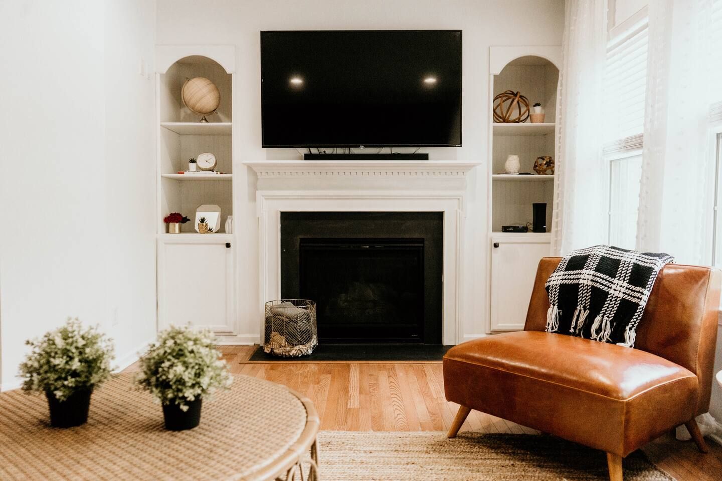 Cozy living room with a wall-mounted black TV above a white fireplace, built-in shelves with decor, a brown leather chair with a black and white plaid throw, and a round woven table.