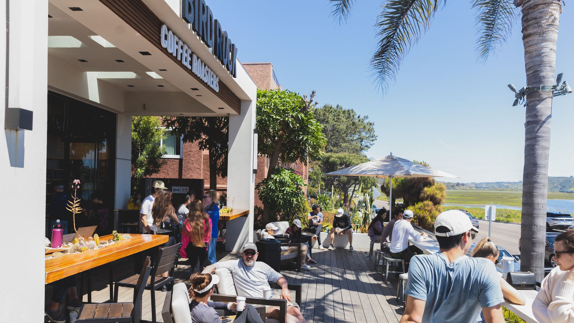 People sit on a patio of a coffee shop overlooking a lagoon on a sunny day.
