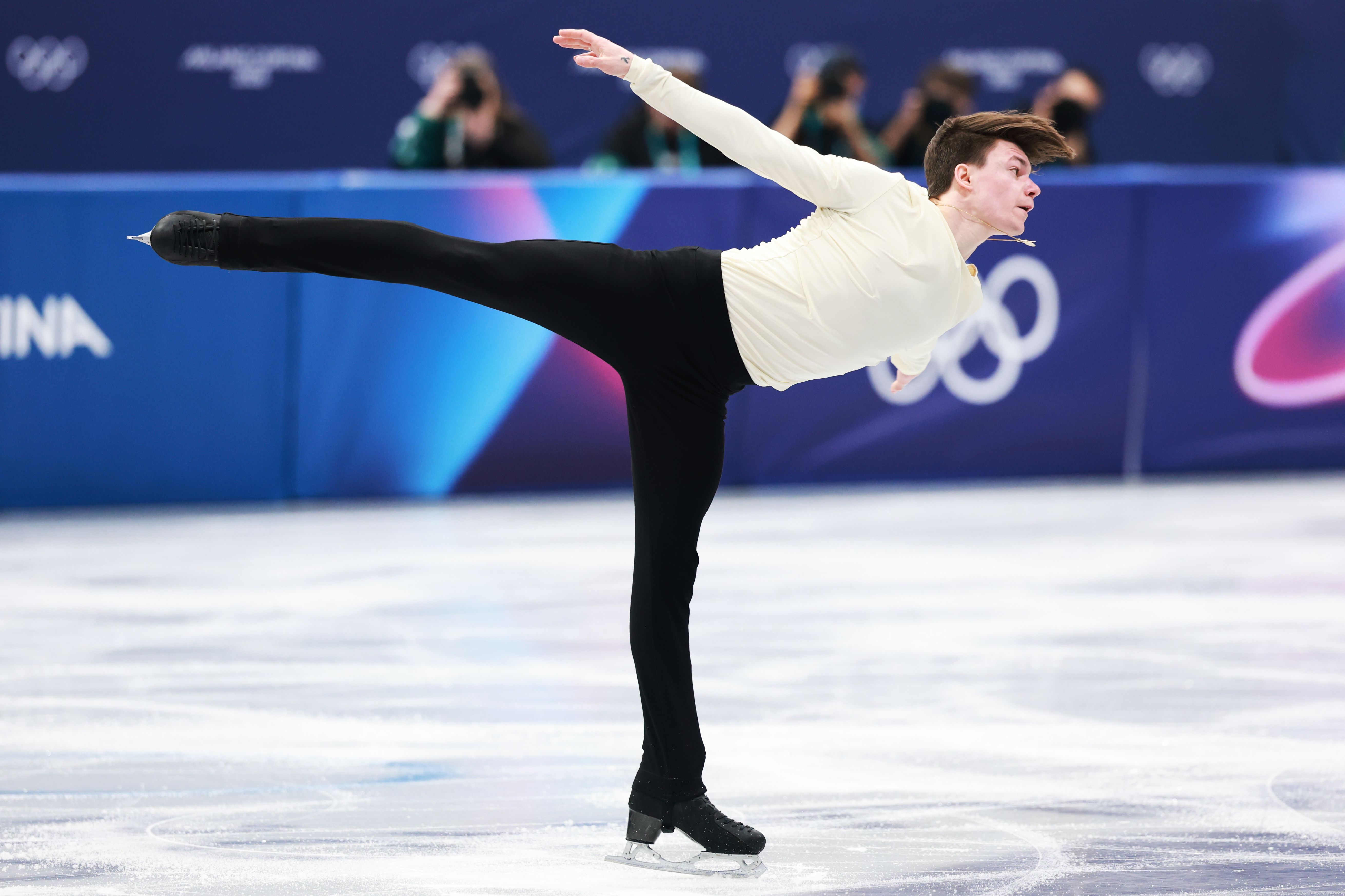 Figure skater Maxim Naumov performing a spiral on ice, wearing black pants and a cream long-sleeve top, with blurred spectators and Olympic rings in the background.