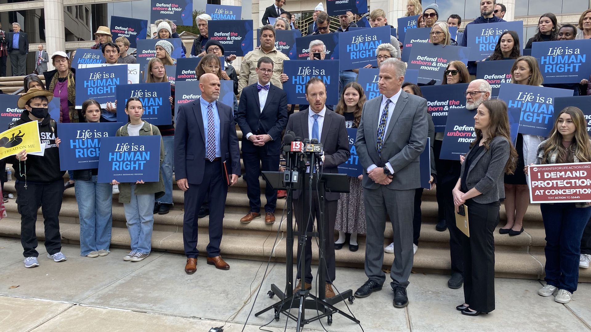 A speaks at a lectern with a group of people on stairs behind him holding signs with anti abortion slogans. 