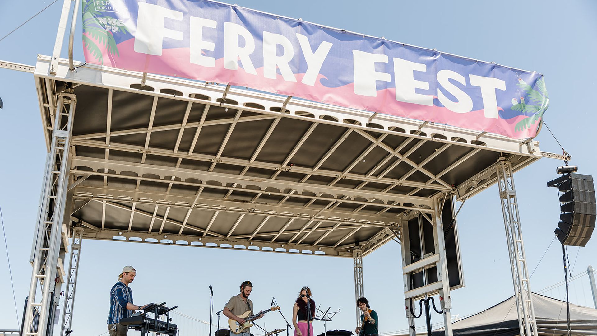 Outdoor stage at Ferry Fest under a blue sky. A banner reads "FERRY FEST" in white on a pink-to-blue gradient; four musicians perform—keyboardist left, guitarist center, two singers, with amps and speakers on stage.