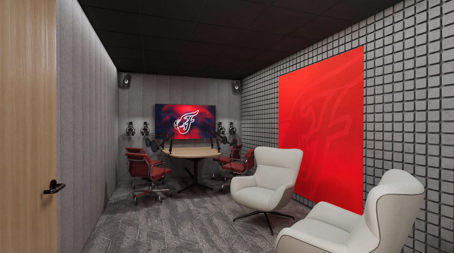 Modern podcast studio: round wooden table with multiple microphones and red chairs around it; gray padded walls, a large red wall panel, white lounge chairs in the foreground, and a screen with a logo.