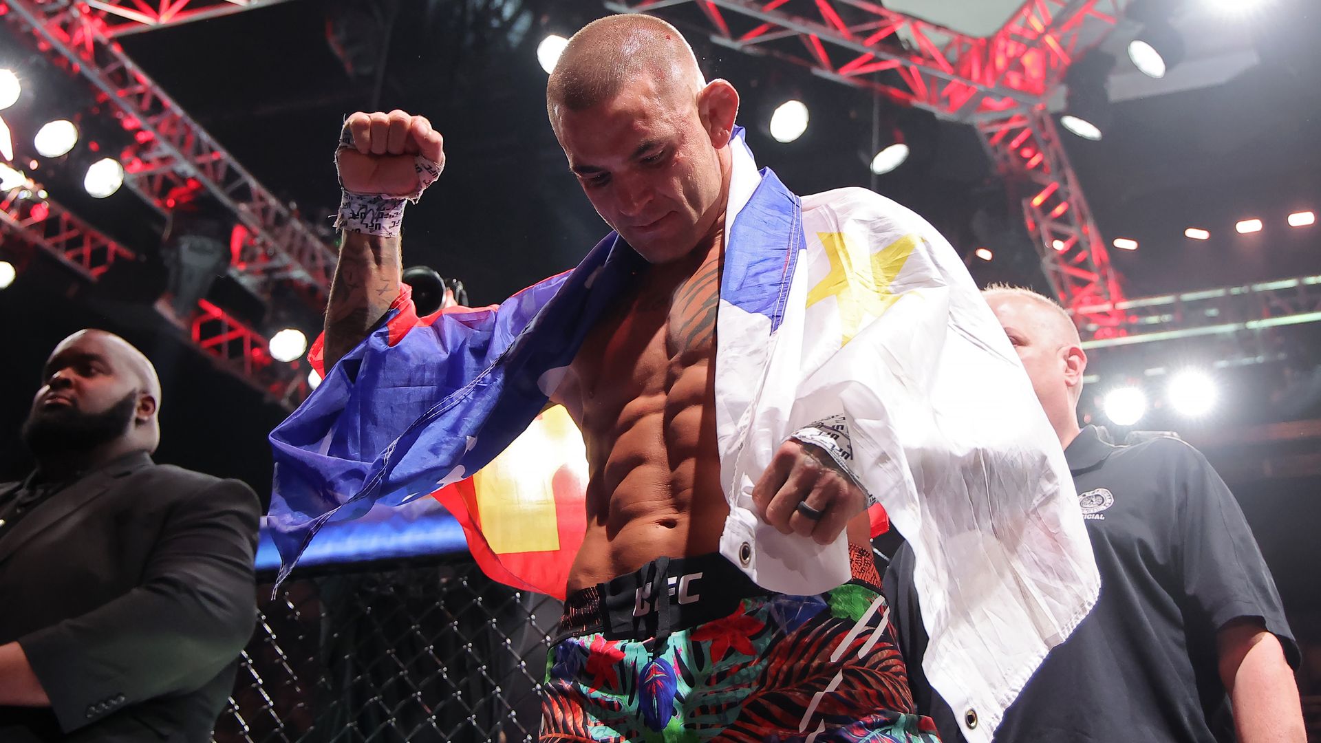 A muscular male UFC fighter with wrapped hands, draped in a blue, red, yellow, and white flag, raises a fist under bright lights with security nearby.