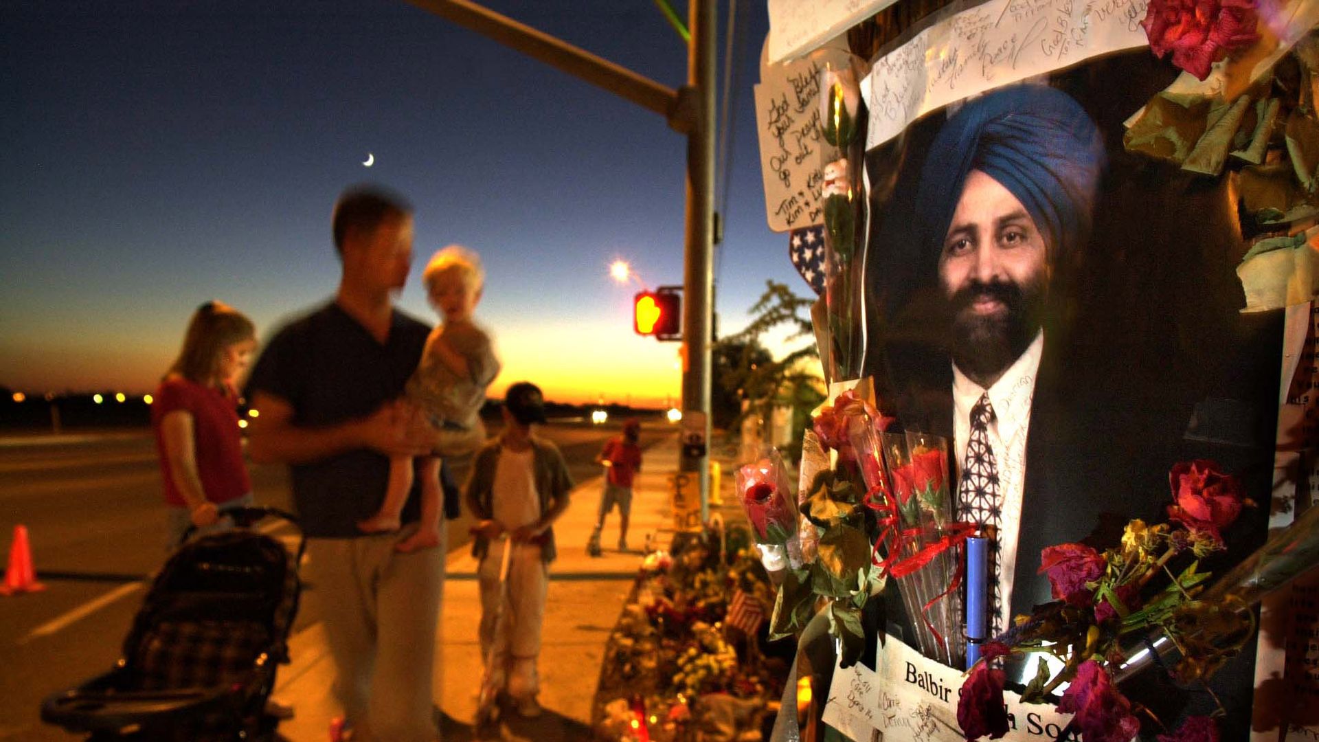 Photo of a picture of Balbir Singh Sodhi plastered on a pole at a memorial site as people look 