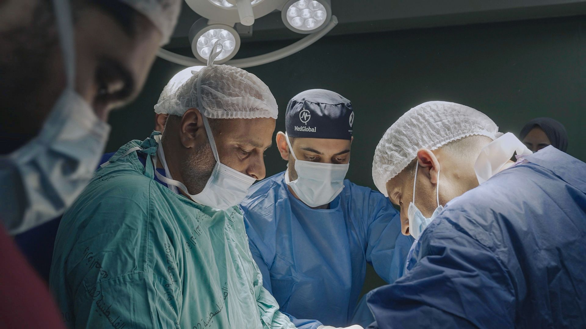 A team of surgeons in scrubs and disposable caps perform surgery under bright overhead lights in an operating room.