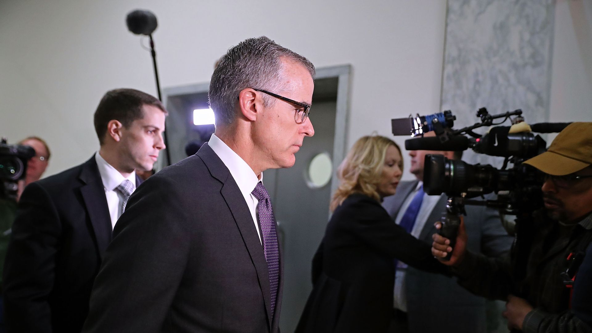 In this image, Andrew McCabe is escorted by a security guard through a light gray hallway in the Rayburn House office building. News cameras follow him. 