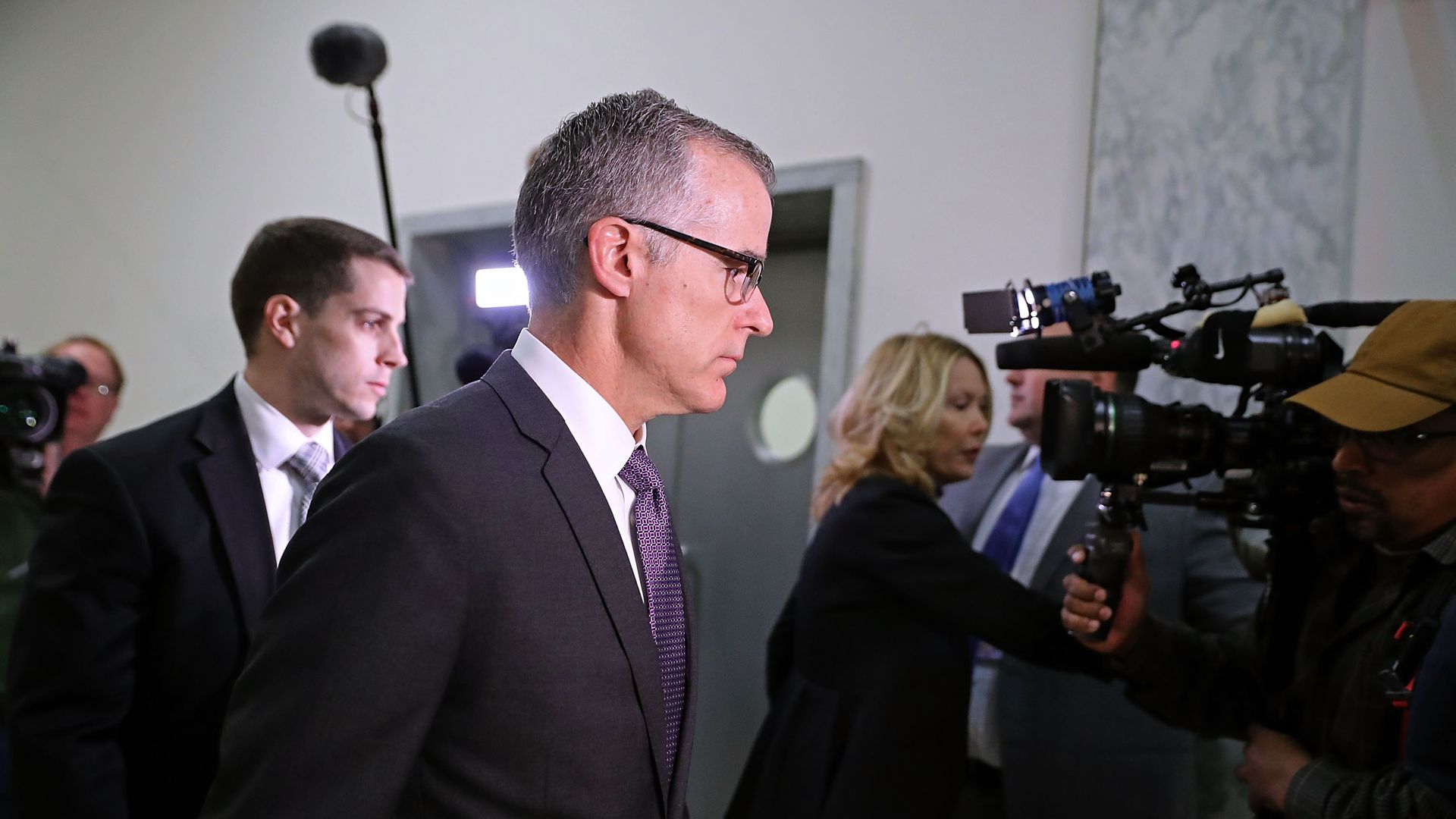 In this image, Andrew McCabe is escorted by a security guard through a light gray hallway in the Rayburn House office building. News cameras follow him.