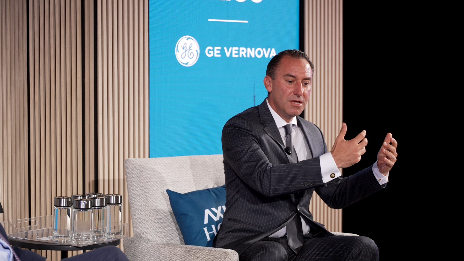 A man in a dark striped suit gestures as he speaks while sitting in a light gray chair with a blue pillow. 