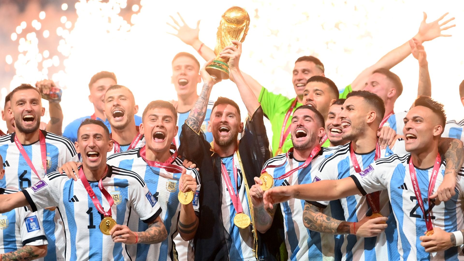 Lionel Messi of Argentina lifts the FIFA World Cup Qatar 2022 Winner's Trophy surrounded by his teammates who are wearing gold medals and shouting and smiling with fireworks in the background.