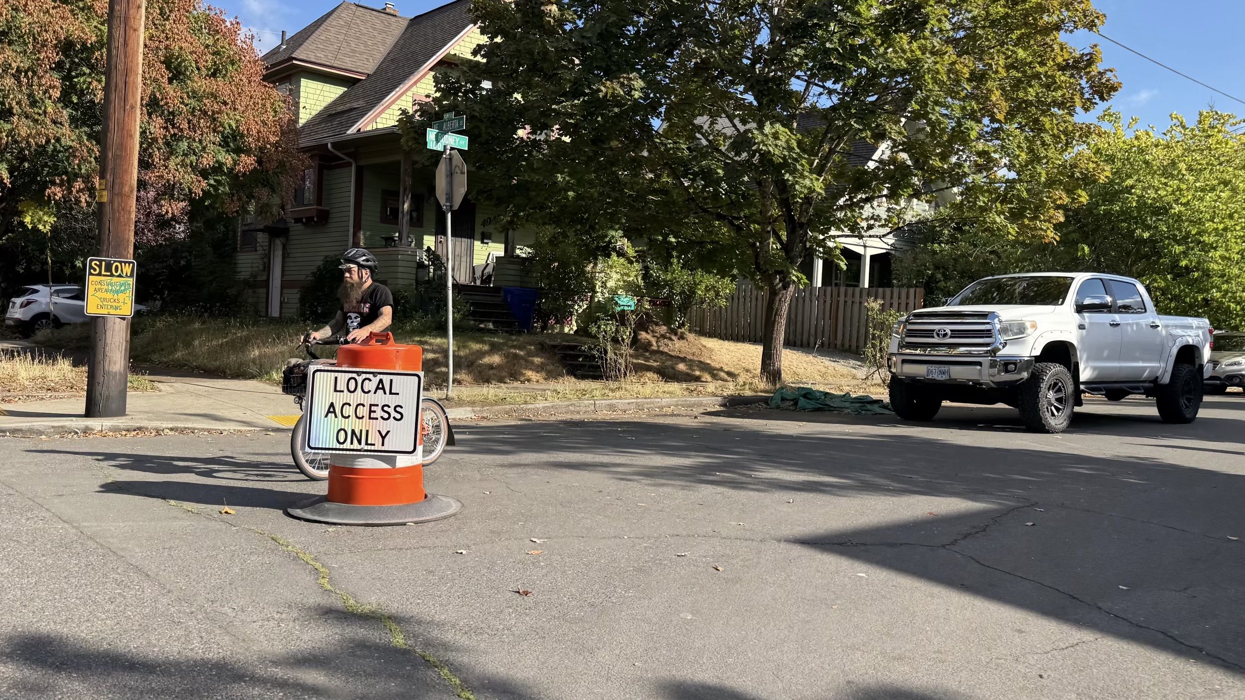 Man wearing a helmet rides a small bike with an orange container on the curb; a cone reads LOCAL ACCESS ONLY. A white pickup sits on the street beside a green house and trees.