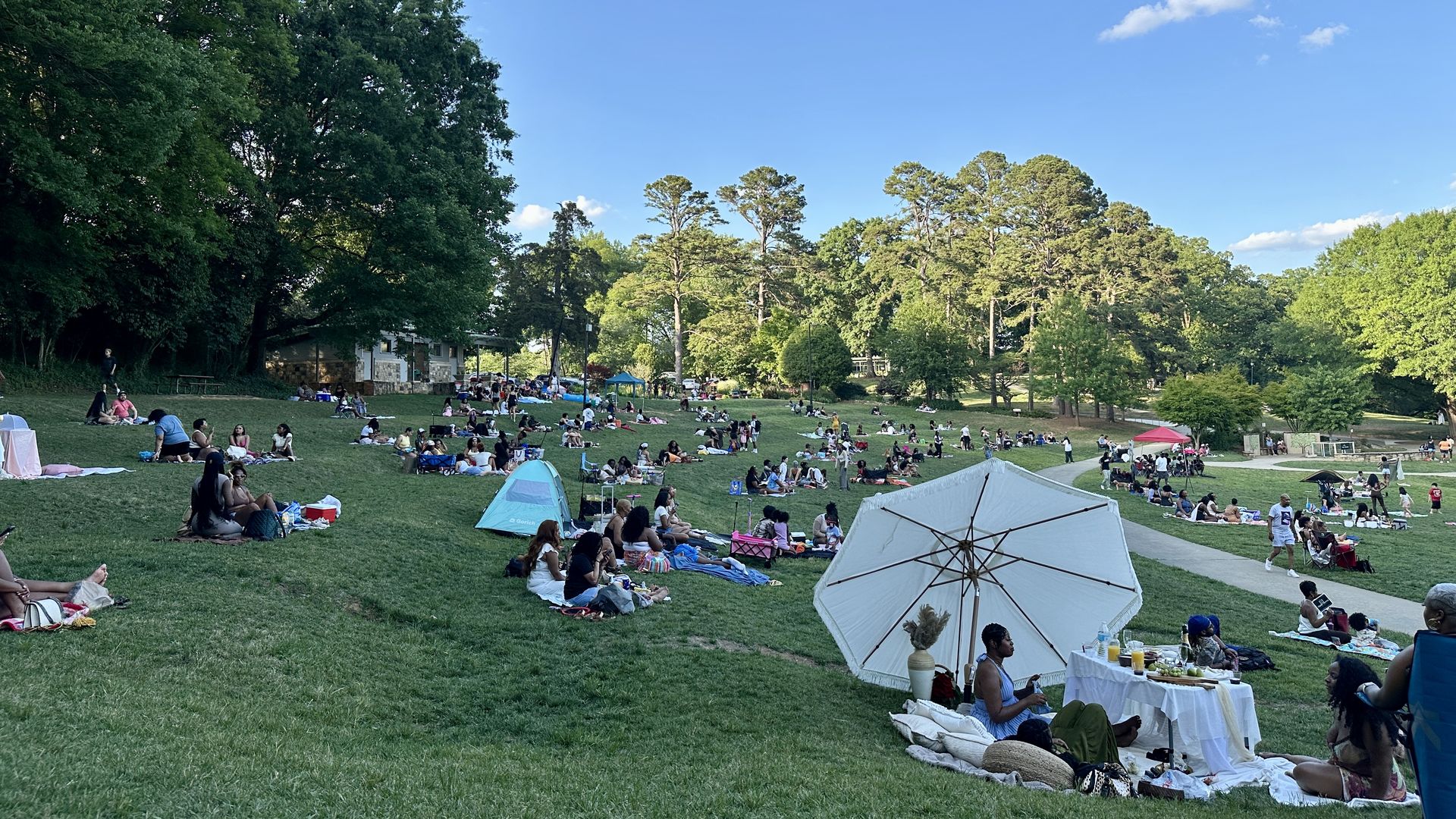 Sunny park with many families picnicking on a grassy lawn near trees. Tents and a white canopy are in the foreground; groups sit on blankets under a clear blue sky with a few clouds.