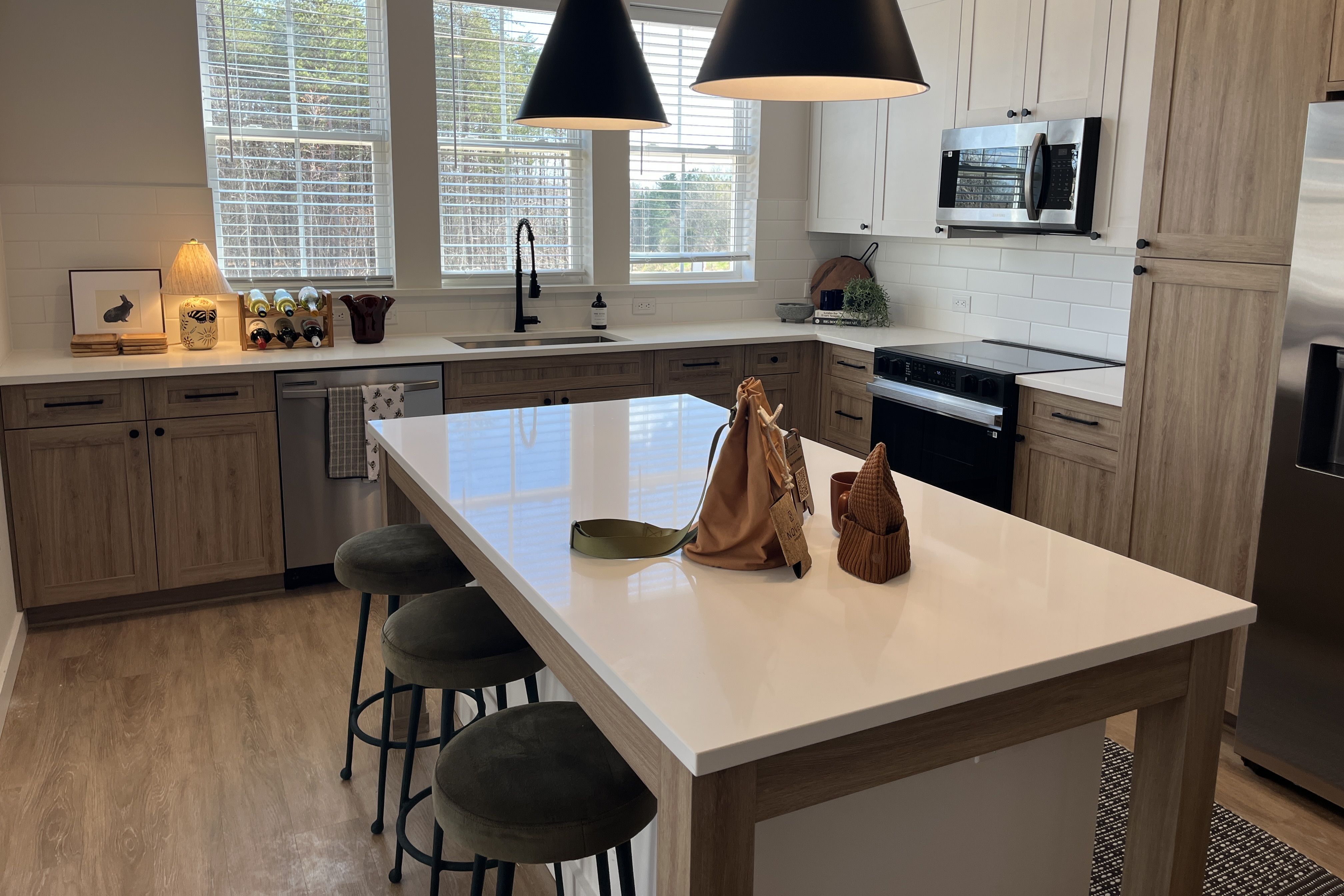 Bright modern kitchen with white countertops, light wood cabinets, and a large island. Black pendant lights, stainless steel appliances, a sink beneath three windows, and green stools.