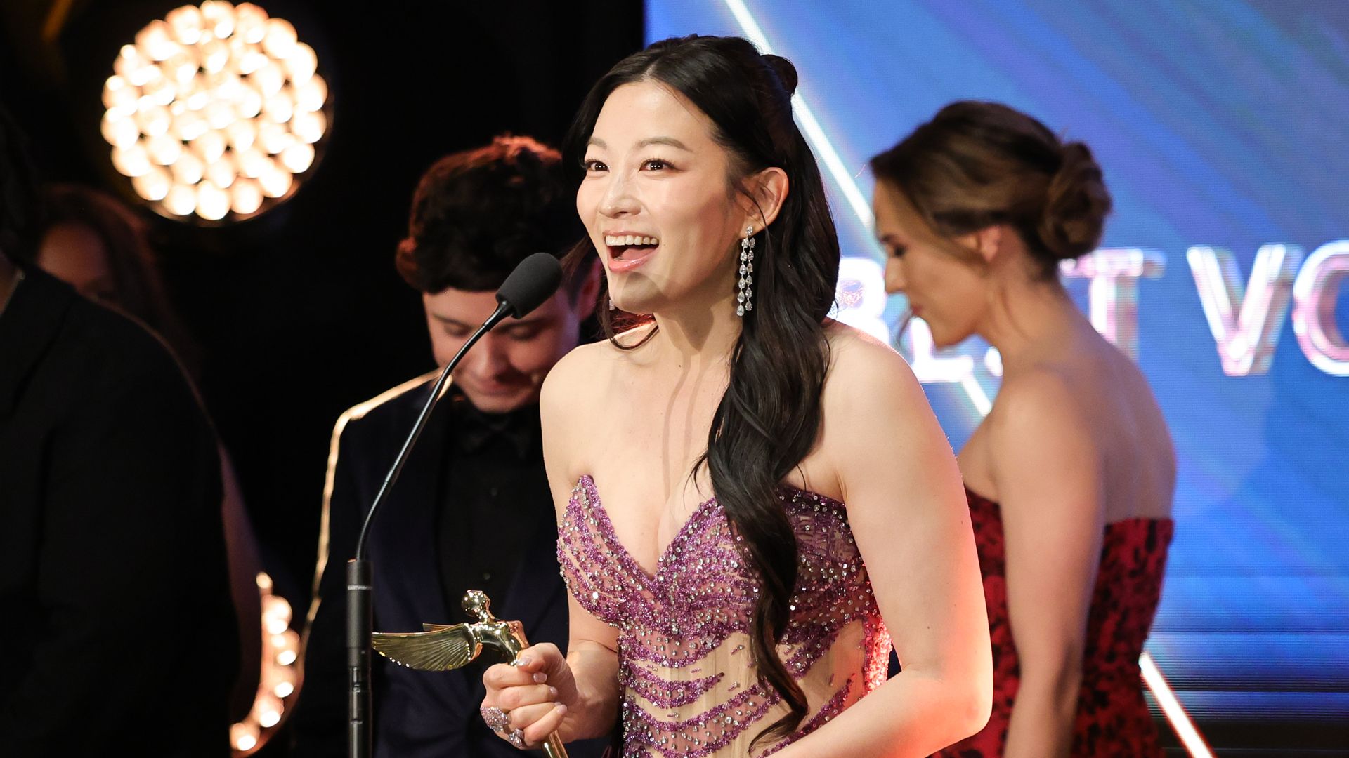 Arden Cho stands on a stage holding a gold award.