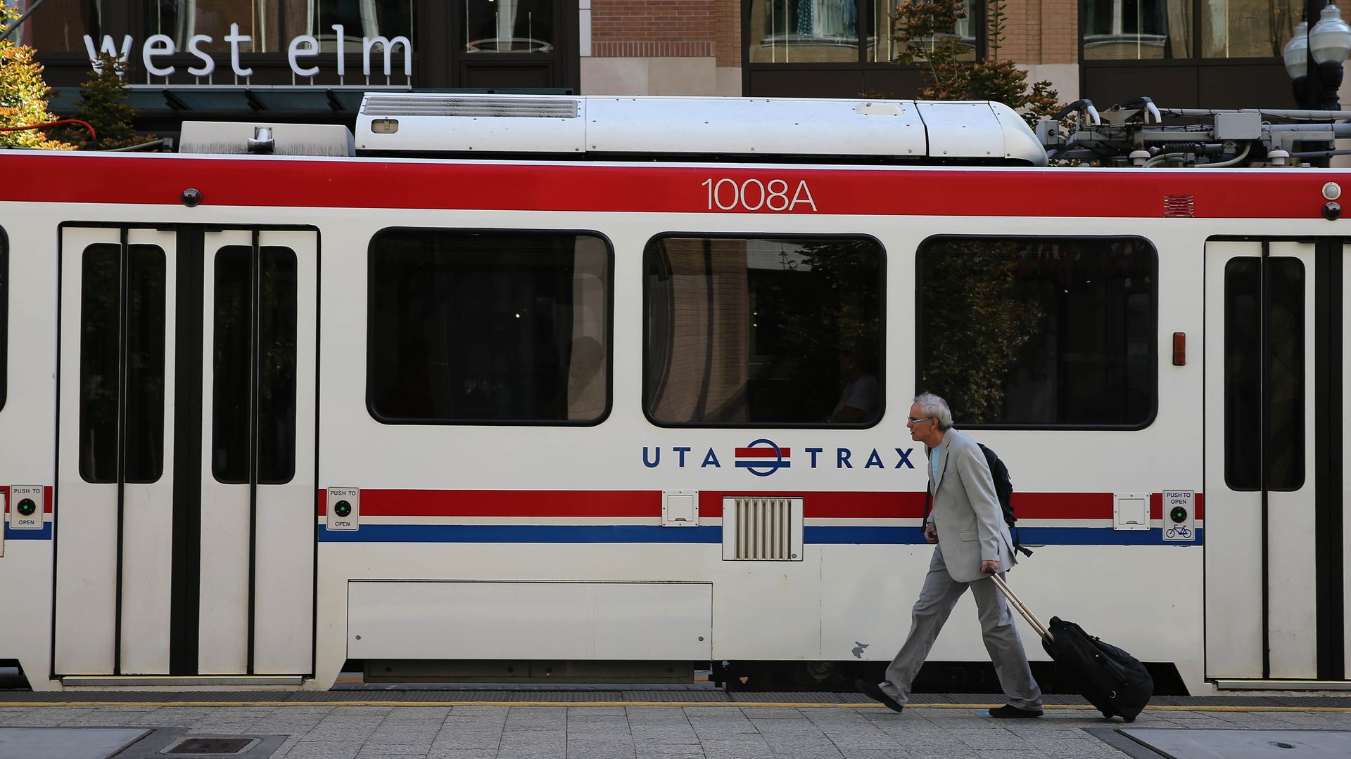 White streetcar with a red stripe, 1008A on its side and UTAH TRAX logo. Behind it, a west elm storefront sign; a man in a gray suit walks by pulling a rolling suitcase.