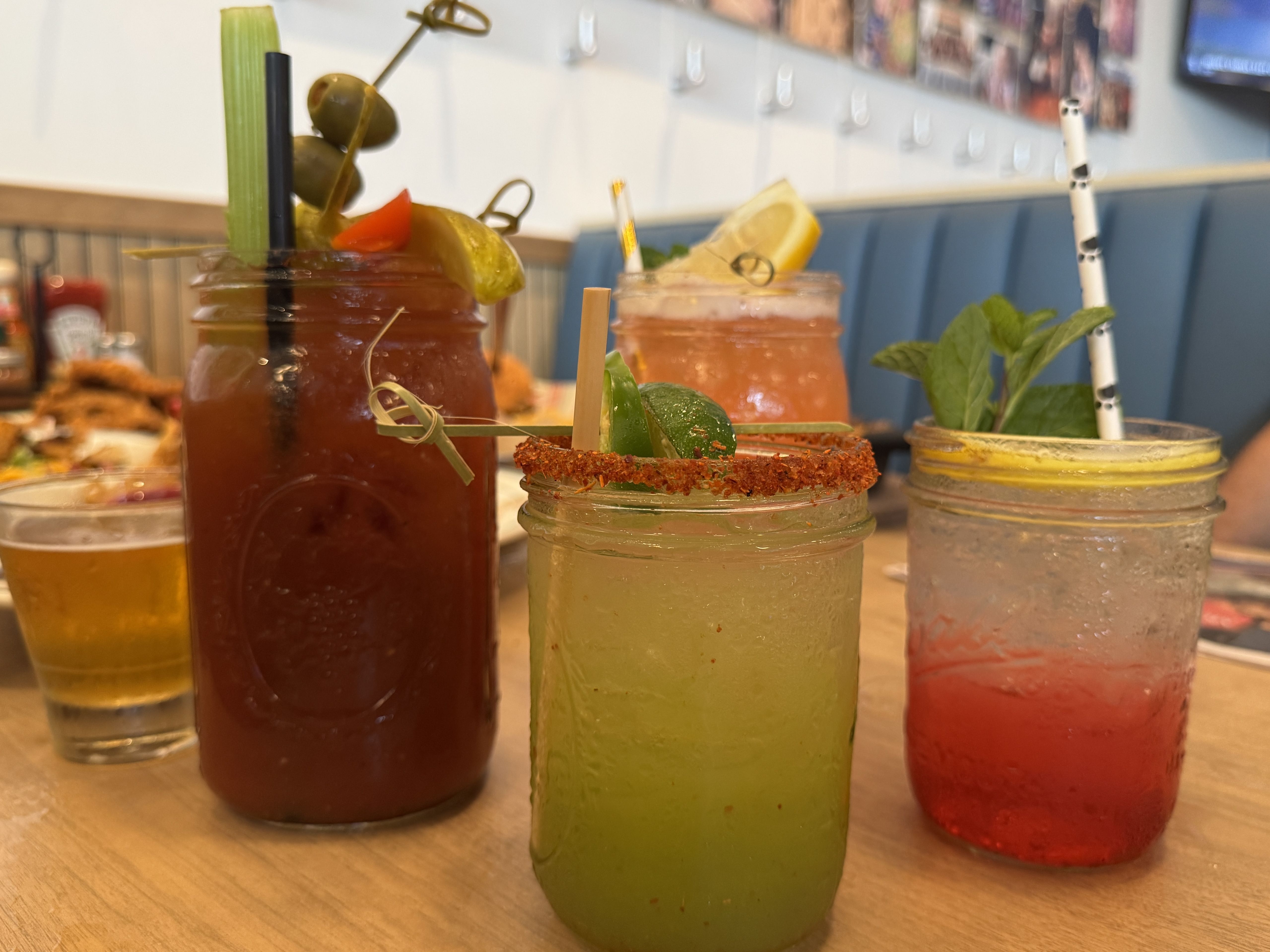 Four colorful drinks in glass jars on a wooden table: a red Bloody Mary with olives and celery, a green drink with chili rim and lime, a pink drink with lemon, and a clear red drink with mint.