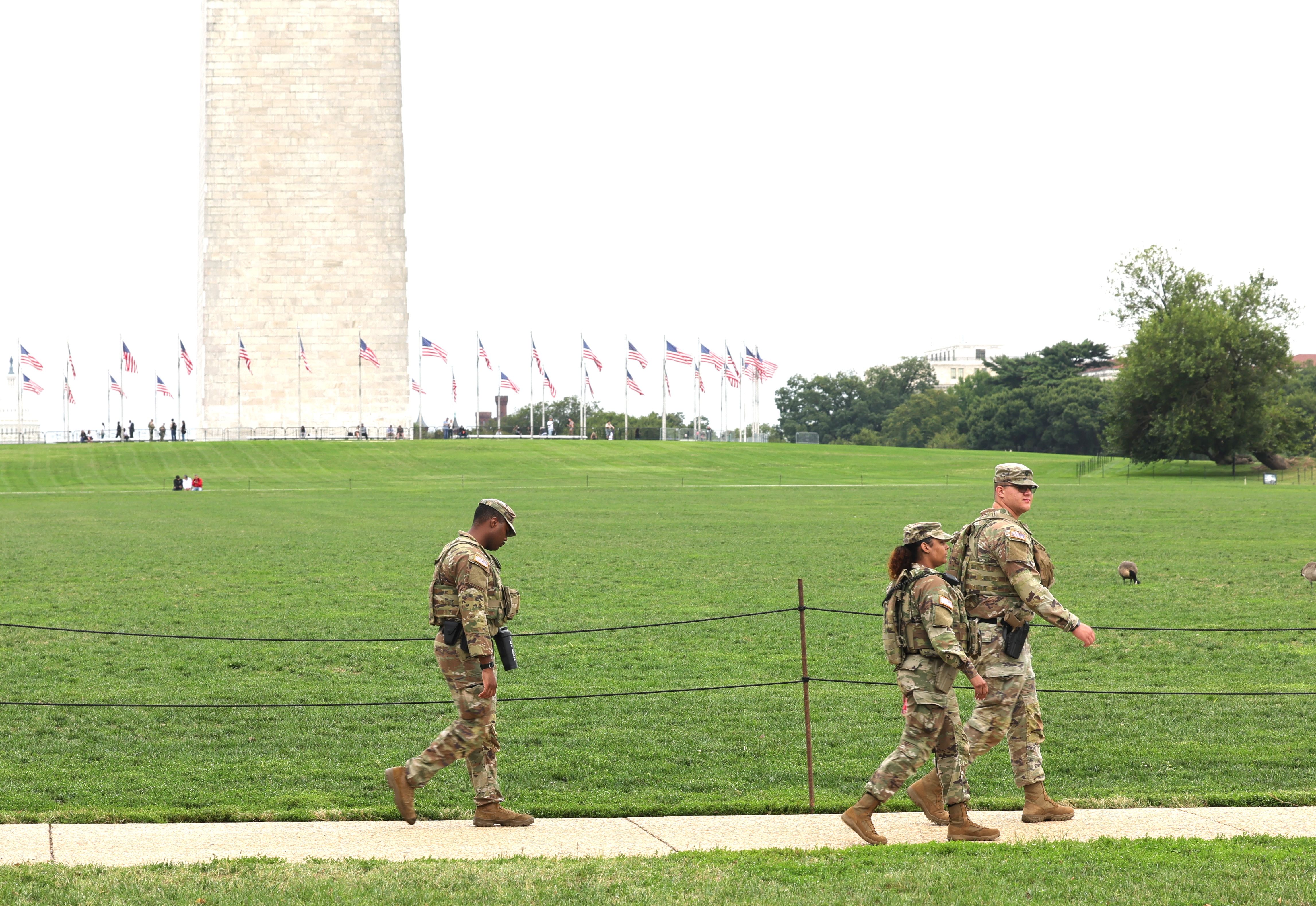 Three U.S. soldiers in camouflage uniforms walking on a path with the Washington Monument and American flags on a green lawn in the background.