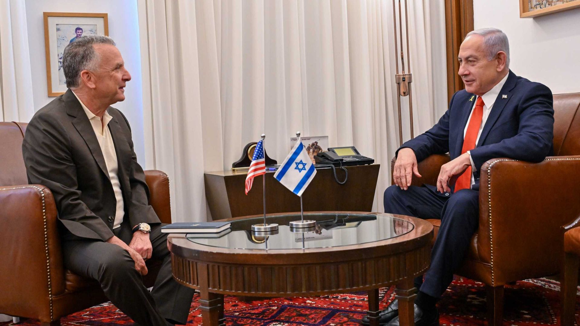 Two men in suits sit across from each other in brown leather chairs with a round glass table between them holding small American and Israeli flags, inside a room with a red patterned carpet.